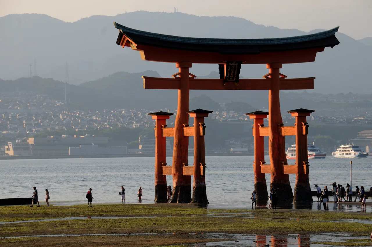 Visitors walking across the exposed seabed to the base of the great torii at low tide, the legs of the gate resting on the sand