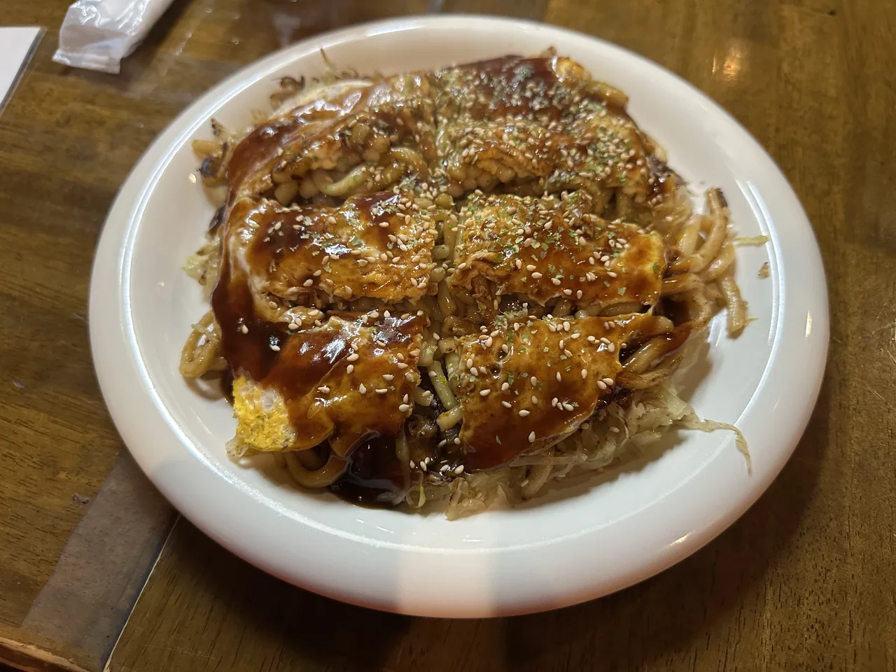 A Hiroshima-style okonomiyaki on a white plate, with noodles visible under the folded egg and brown sauce, topped with sesame seeds