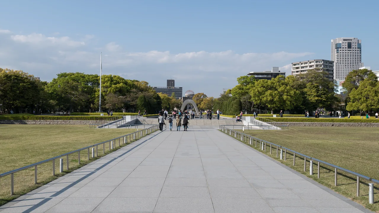 A wide paved walkway through Hiroshima Peace Memorial Park leading towards the arched cenotaph, with the A-Bomb Dome visible beyond it