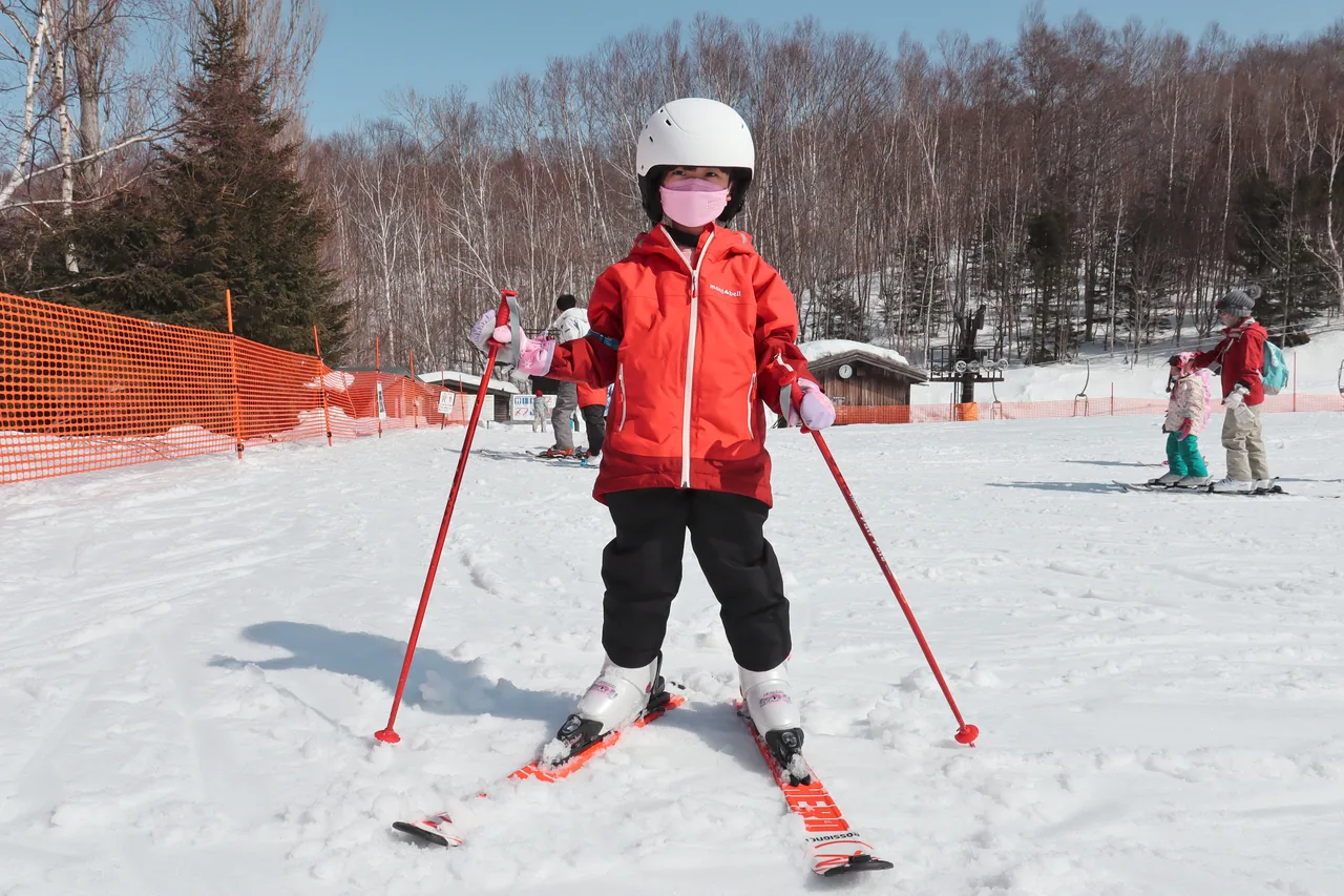 A child in a helmet and red jacket standing on skis holding poles, facing the camera on a sunny day, with orange safety fencing, other children and an instructor in the ski-school area behind