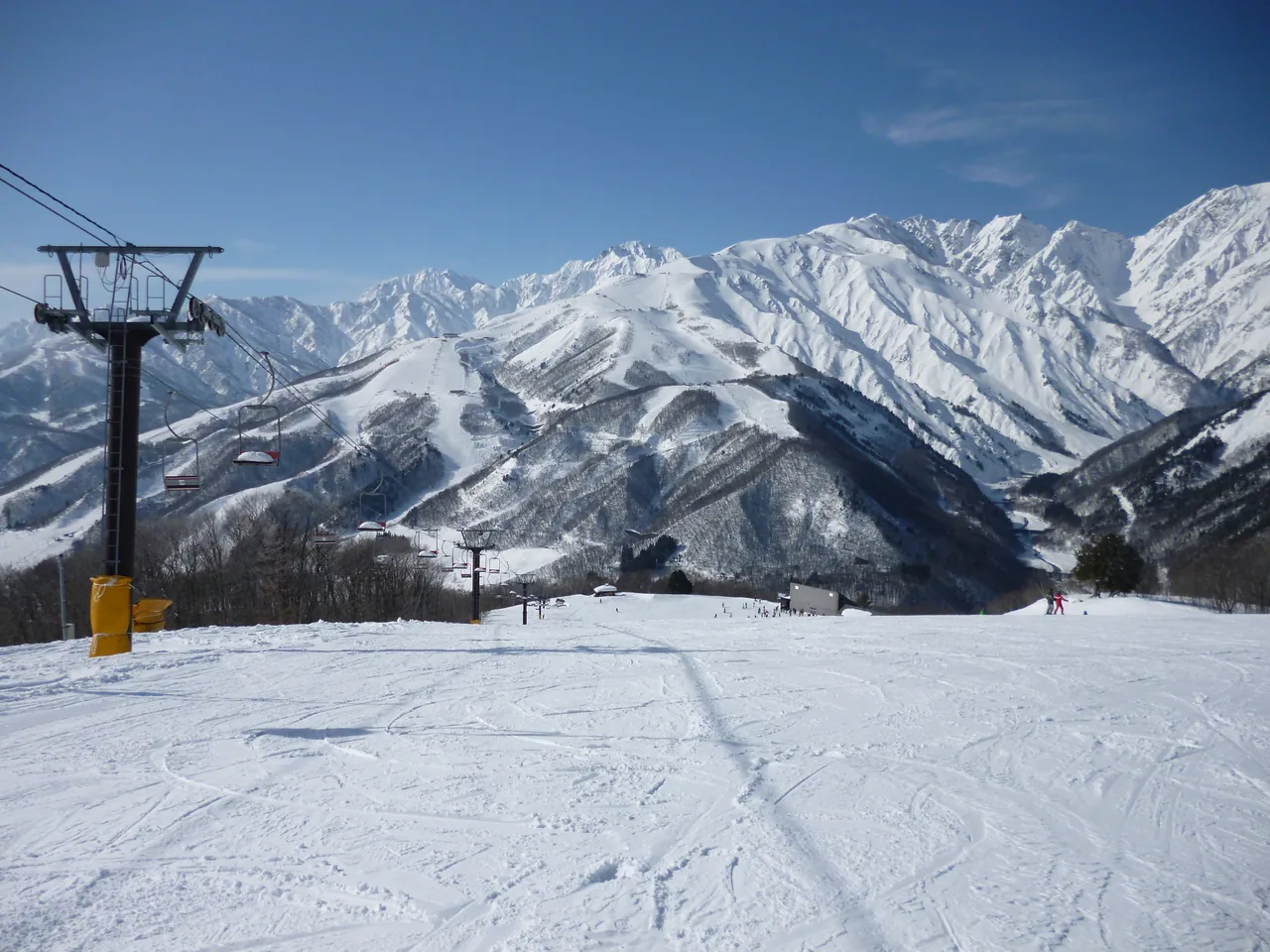 A wide groomed piste at Hakuba Iwatake in Nagano, with a chairlift tower and empty chairs on the left, the snow-covered Hakuba range and deep blue sky behind, and two or three skiers far off to the right