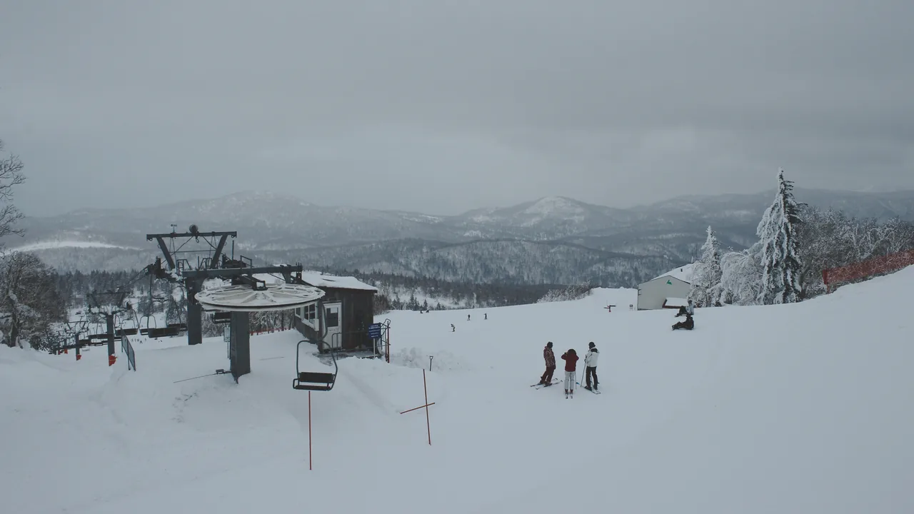 The top station of a lift at Sapporo Kokusai with chairs on the left, three skiers standing on the gentle groomed run beside it and snow-covered ridges in the distance, under flat overcast light