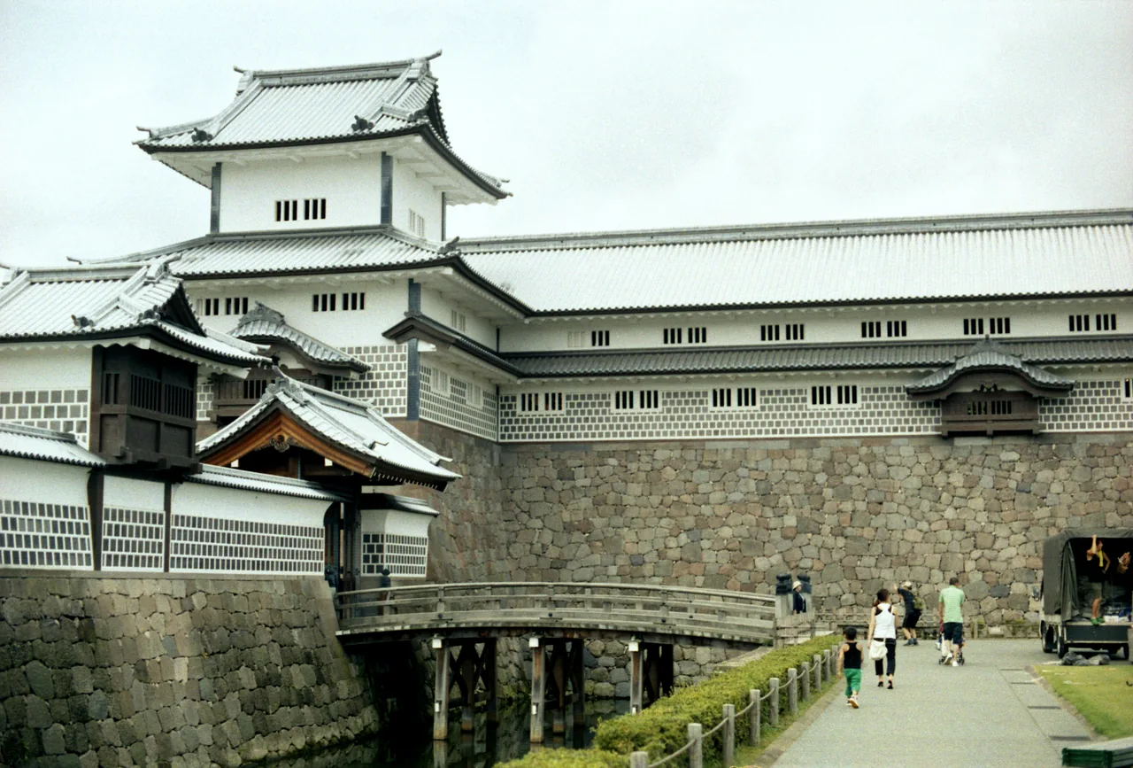 The long white walled Gojukken Nagaya storehouse and the Hishi Yagura turret of Kanazawa Castle above a stone base, with a wooden bridge in front