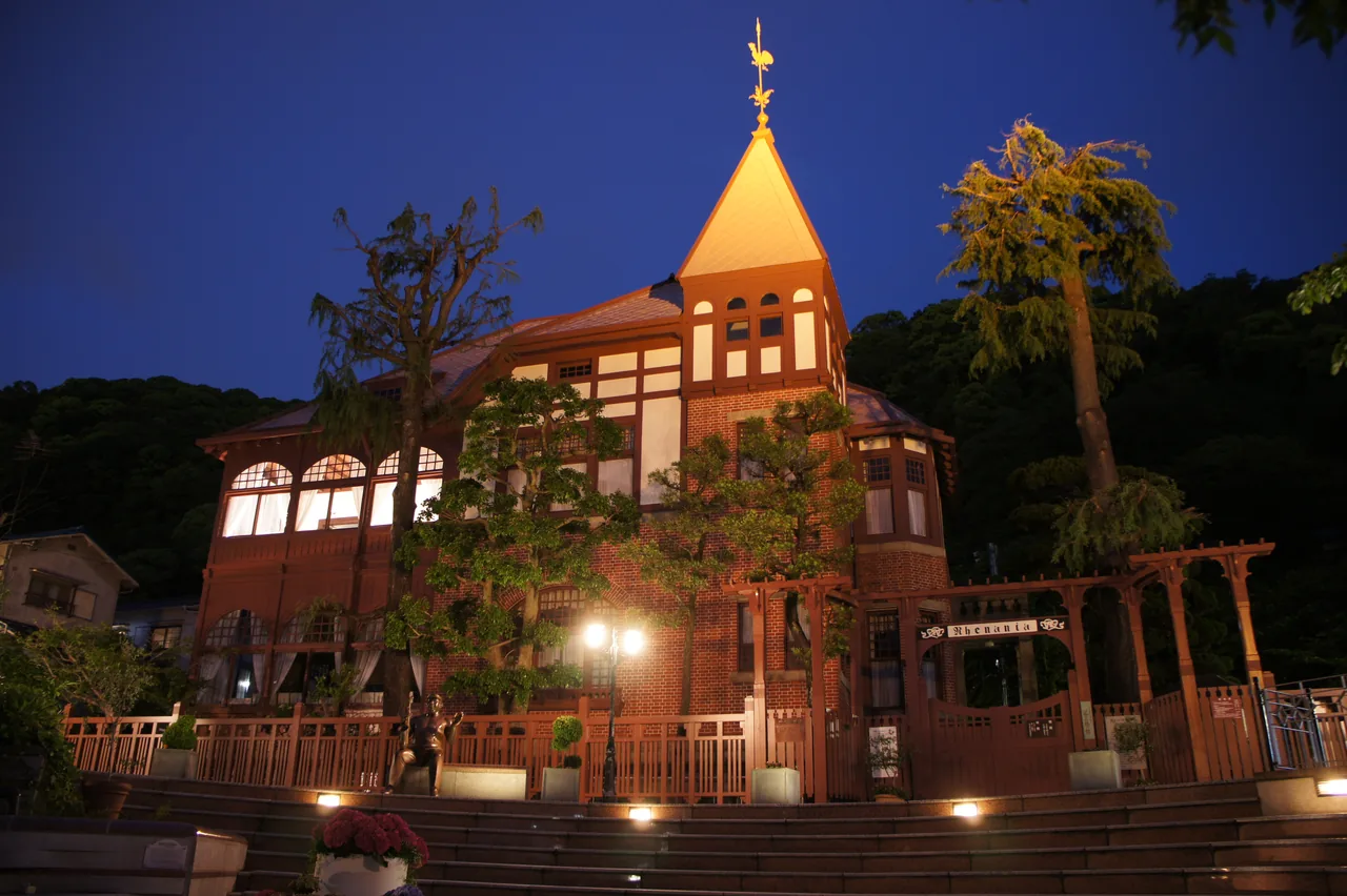 The Weathercock House in Kitano lit up at night, its brick facade and pointed turret standing above a stone terrace
