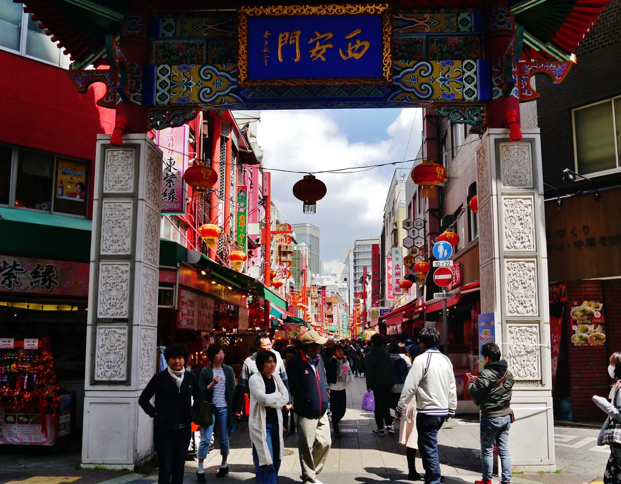 The ornate gate of Kobe Chinatown with red lanterns strung down the street beyond it and people walking through