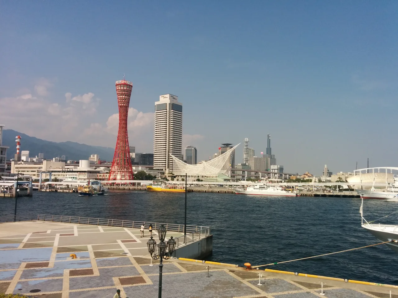 The red lattice of Kobe Port Tower seen across the harbour from Meriken Park, with the city and the Rokko ridge behind it