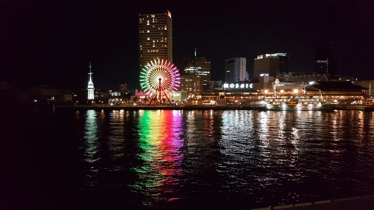 The lit ferris wheel and the Mosaic building at Kobe Harborland after dark, their colours running down the water in long reflections