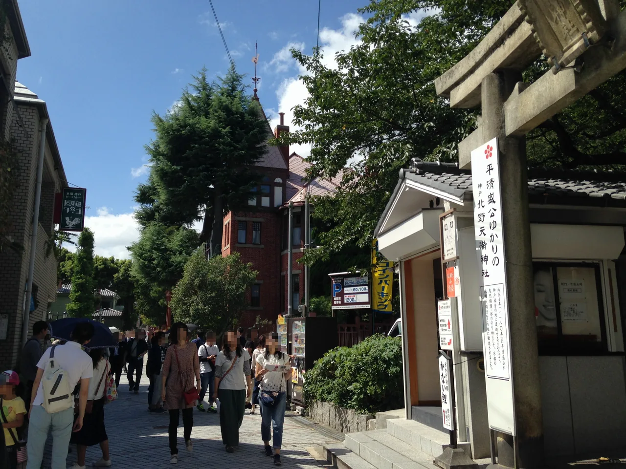 A busy street in the Kitano foreign residence district, with the red brick Weathercock House on the left, a shrine torii on the right and people walking up the slope