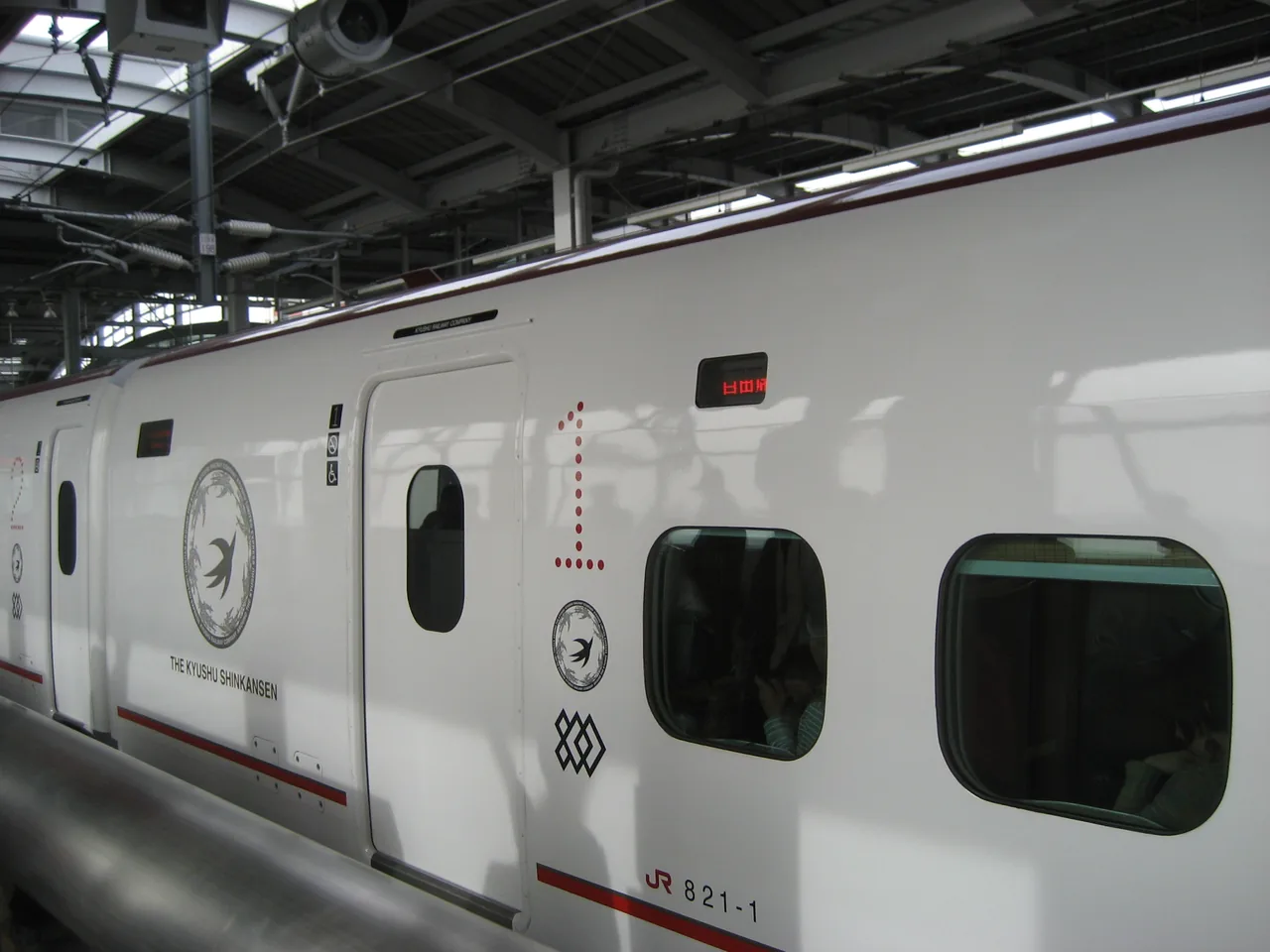 The side of an 800 series Kyushu Shinkansen car at a platform, with round windows and a dark band