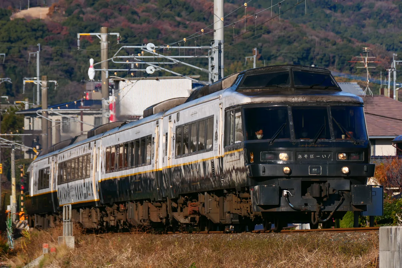 The dark blue Aso Boy train running on the line with wooded hills behind