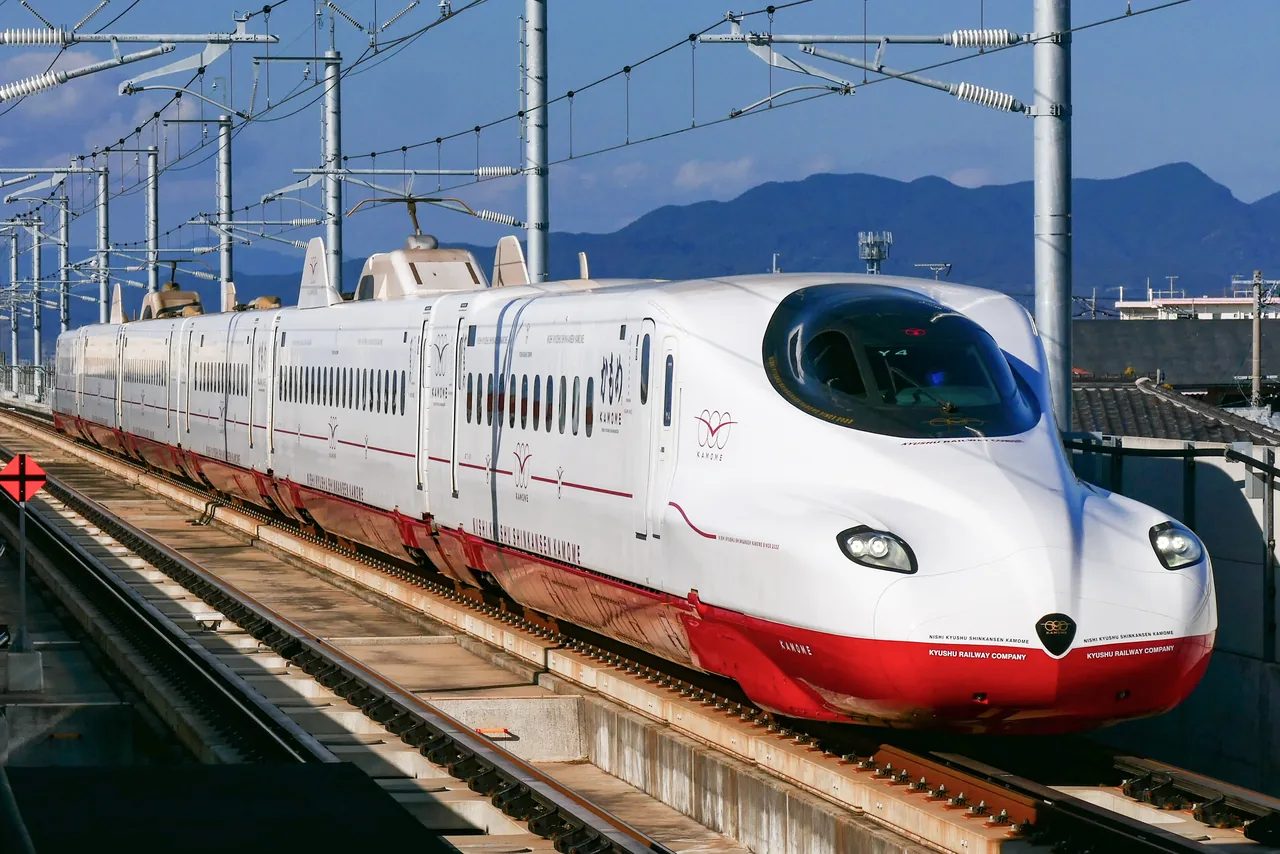 A white and red N700S Kamome train of the Nishi Kyushu Shinkansen standing at a platform with hills behind it
