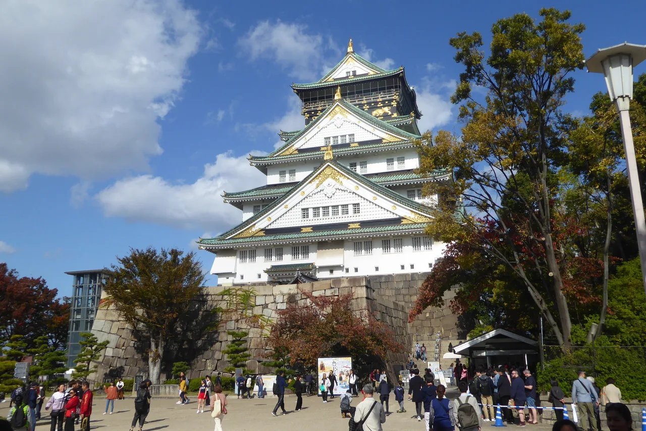 Osaka Castle keep under a blue sky with visitors crossing the plaza in front of it