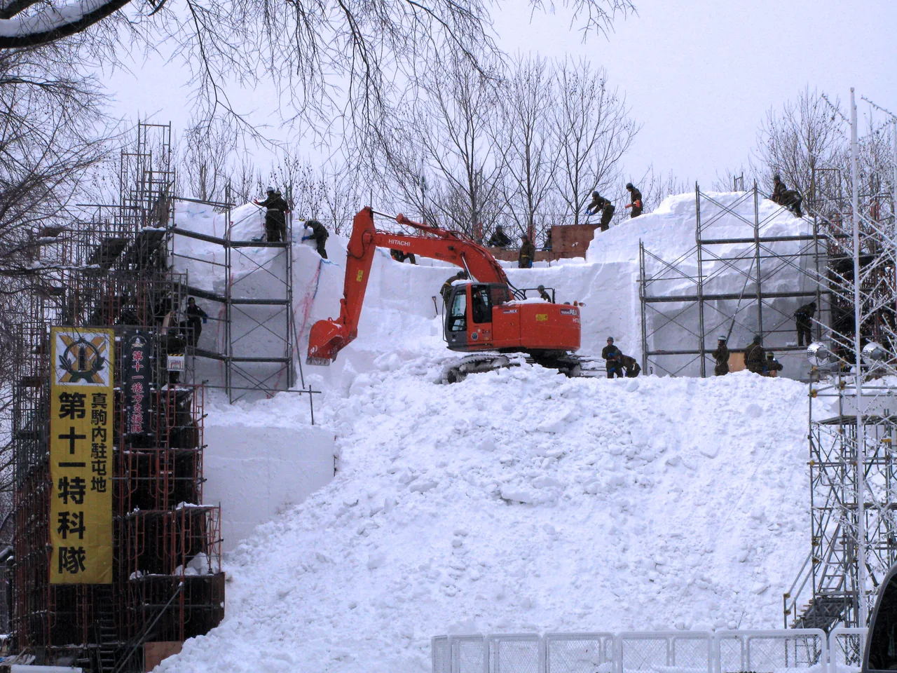 An excavator piling snow inside a construction frame at the festival site