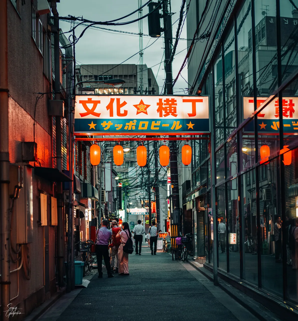 A narrow Sendai alley under a red Bunka Yokocho sign with red lanterns hung along it and people walking through