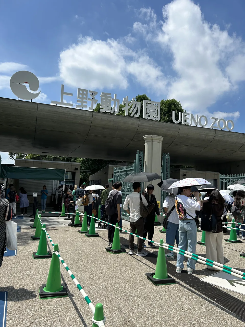 A queue of visitors under the Ueno Zoo entrance sign, several holding umbrellas against the sun