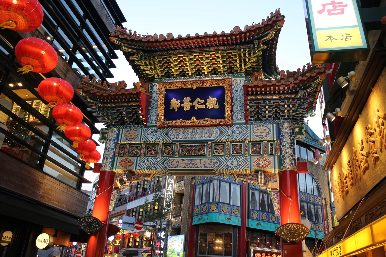 A gate of Yokohama Chinatown seen from the street, painted in colour with a tiled roof, with shopfronts and pedestrians beyond it