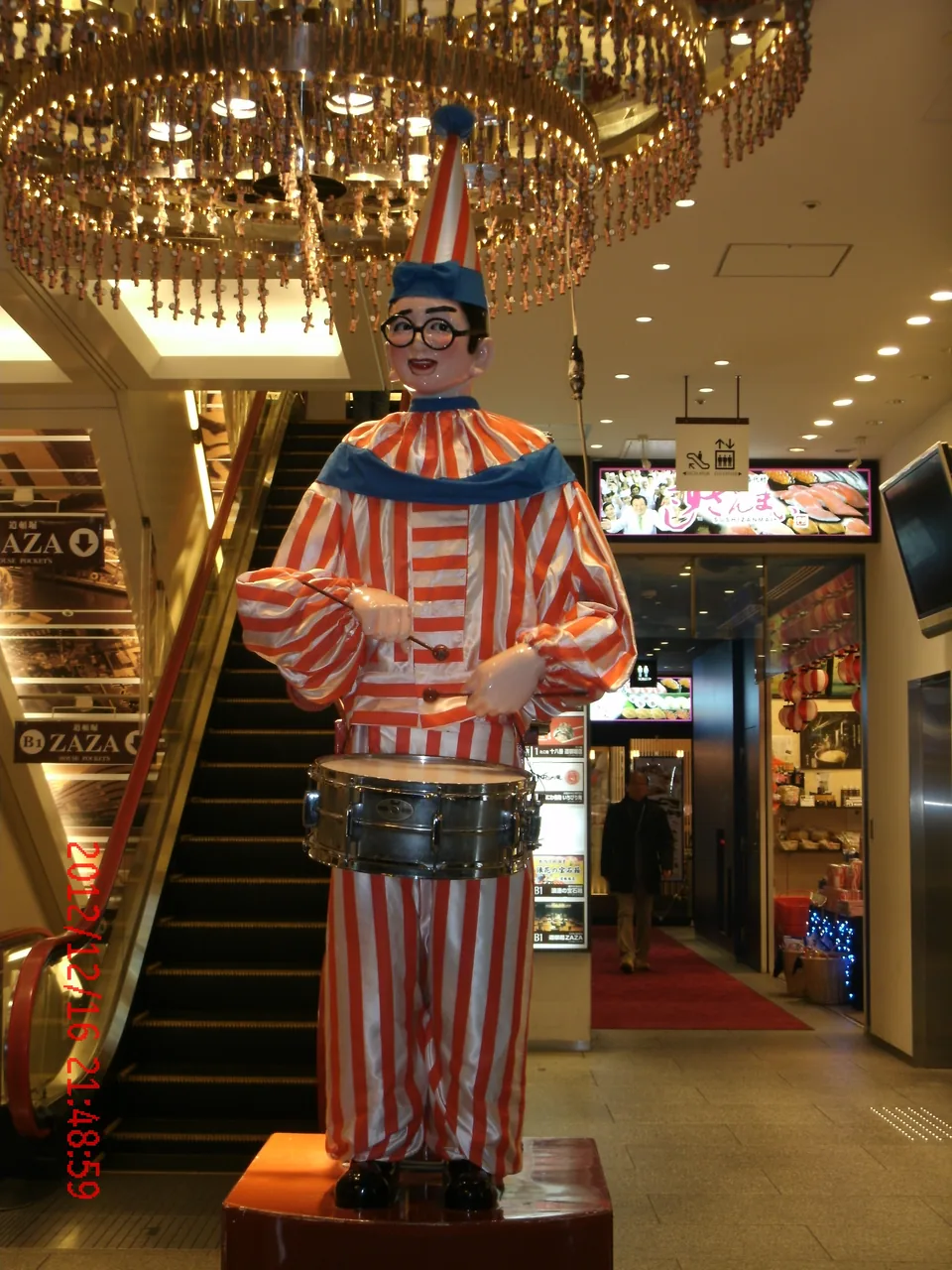 A figure in a striped outfit standing with a drum at a building entrance