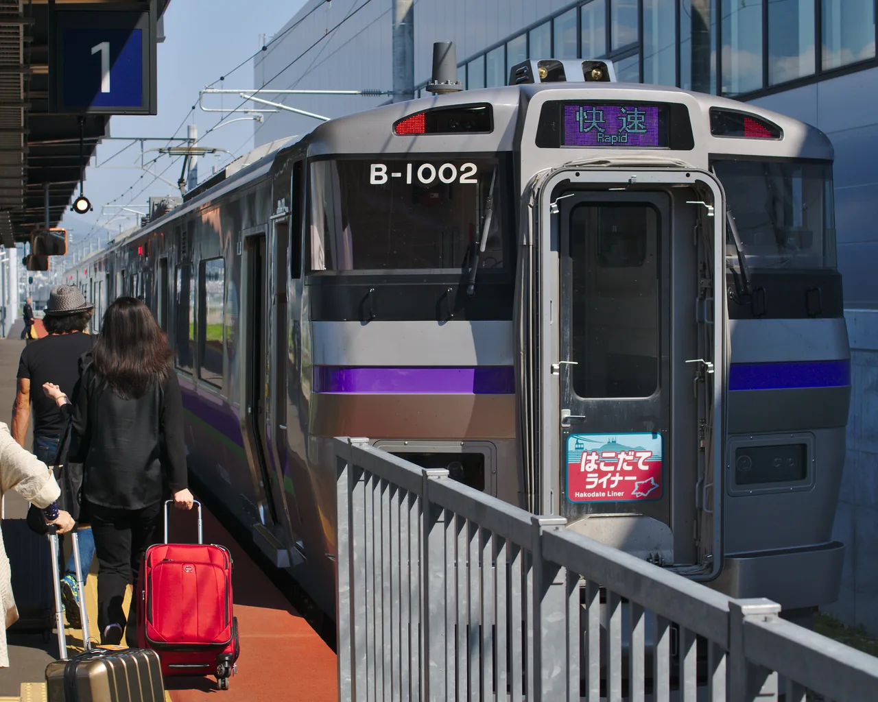 A commuter train at a platform with a passenger and a suitcase beside it