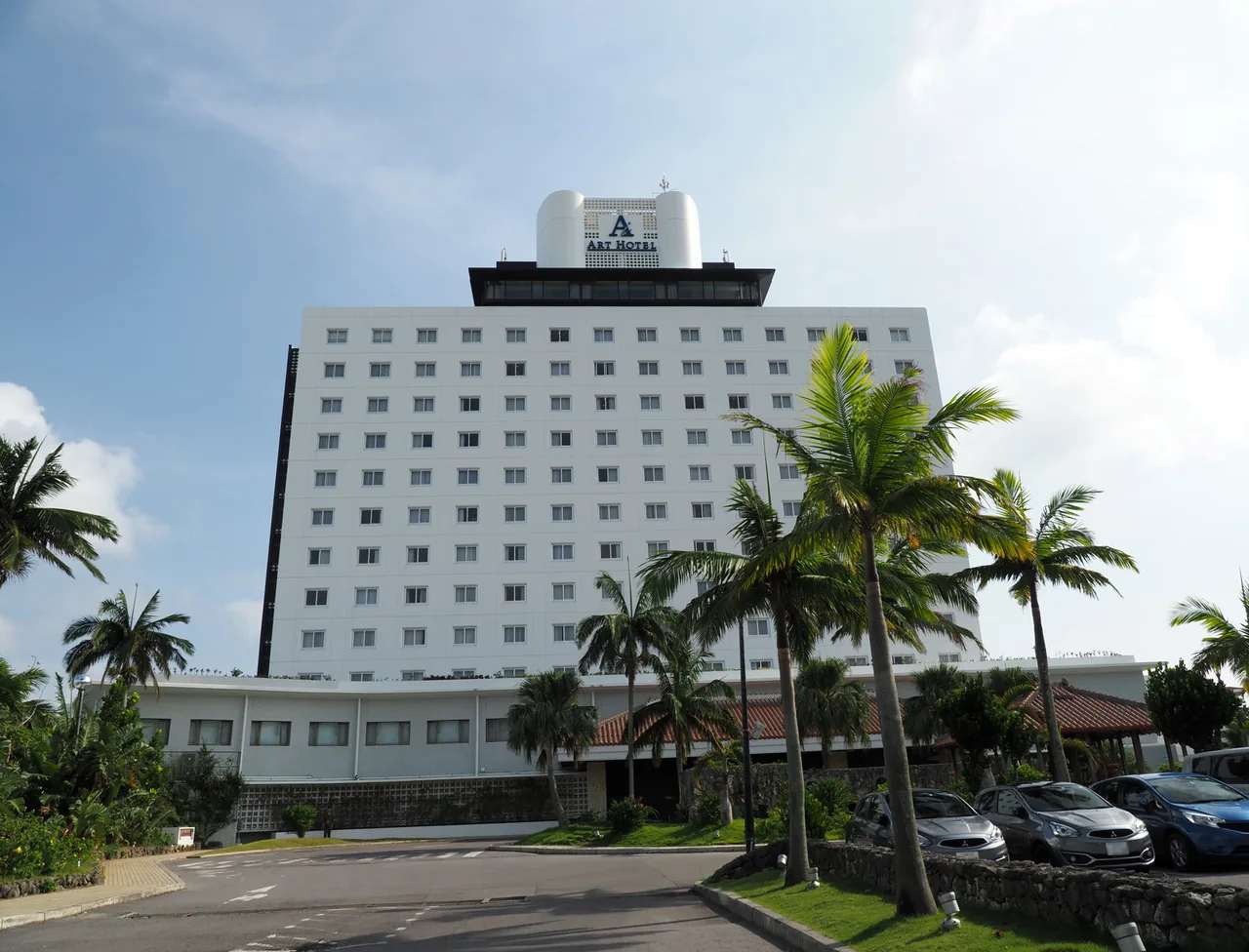 A tall white hotel building behind palm trees and a car park in Ishigaki town