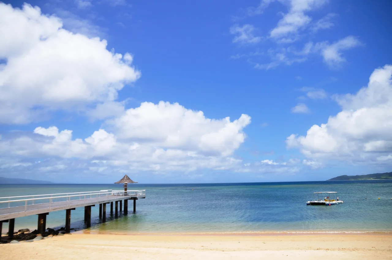 A wooden pier running out over shallow water from a white sand beach under a blue sky