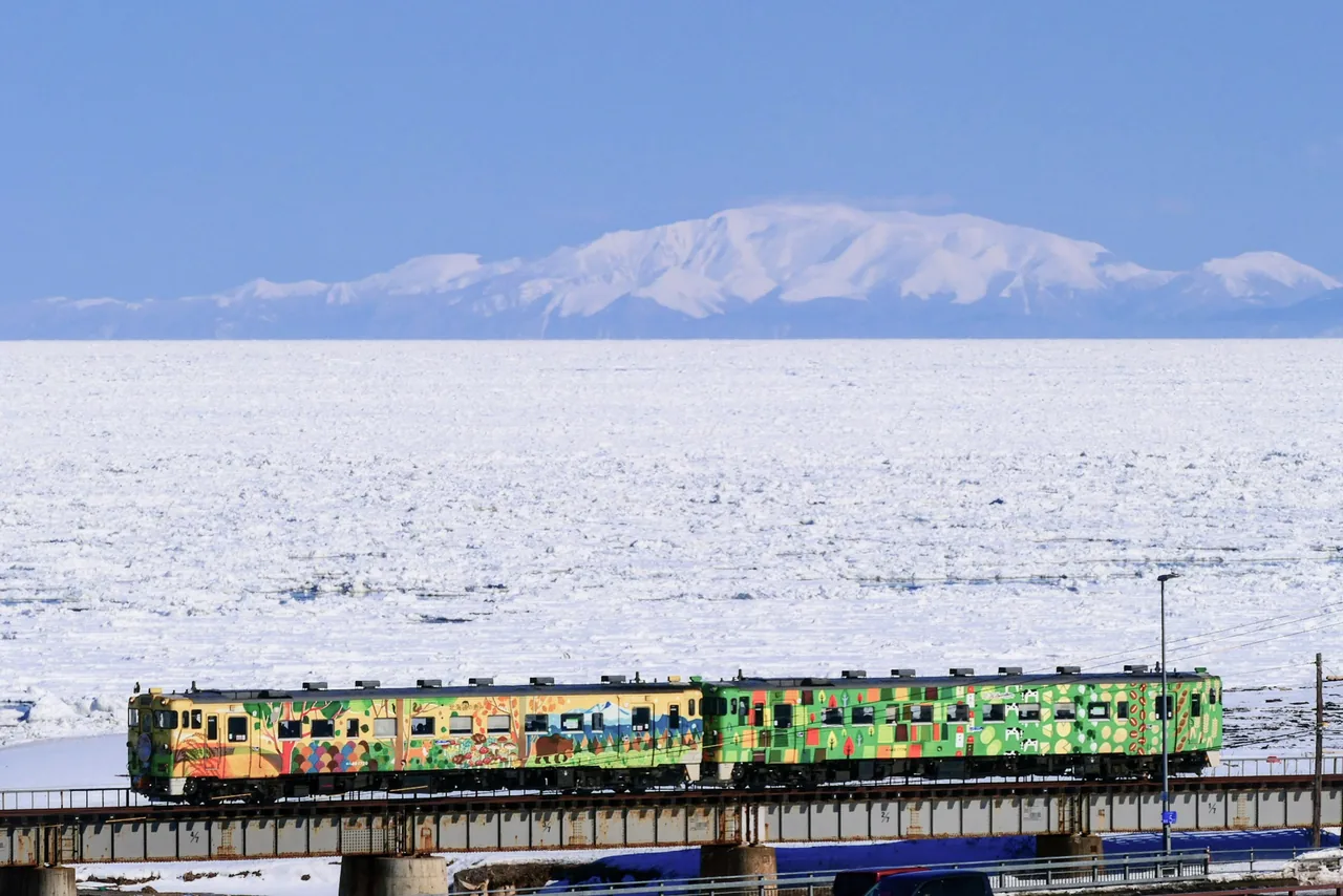 A single-car diesel train crossing a coastal bridge, with the sea covered in white drift ice and snowy mountains behind