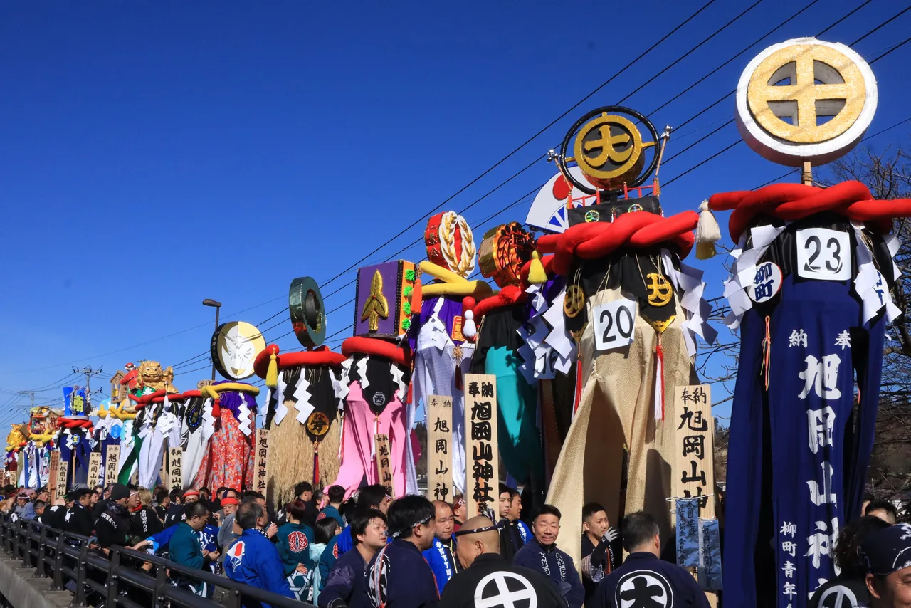 A procession carrying tall, colourful decorated poles across a snowy bridge under a clear sky