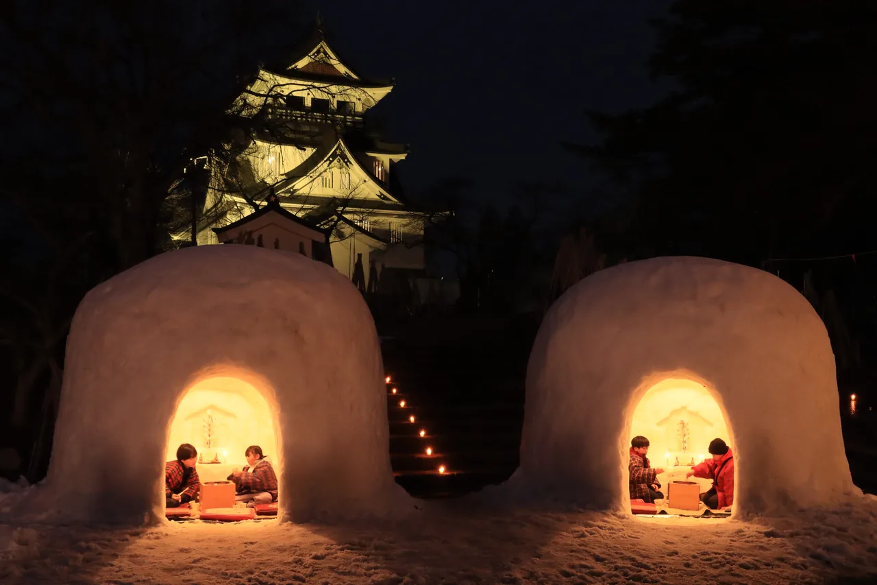 Two small snow huts lit from inside at night, with the illuminated Yokote Castle behind