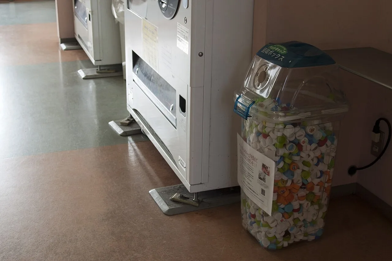 A clear collection box beside a vending machine, filled to the top with plastic bottle caps