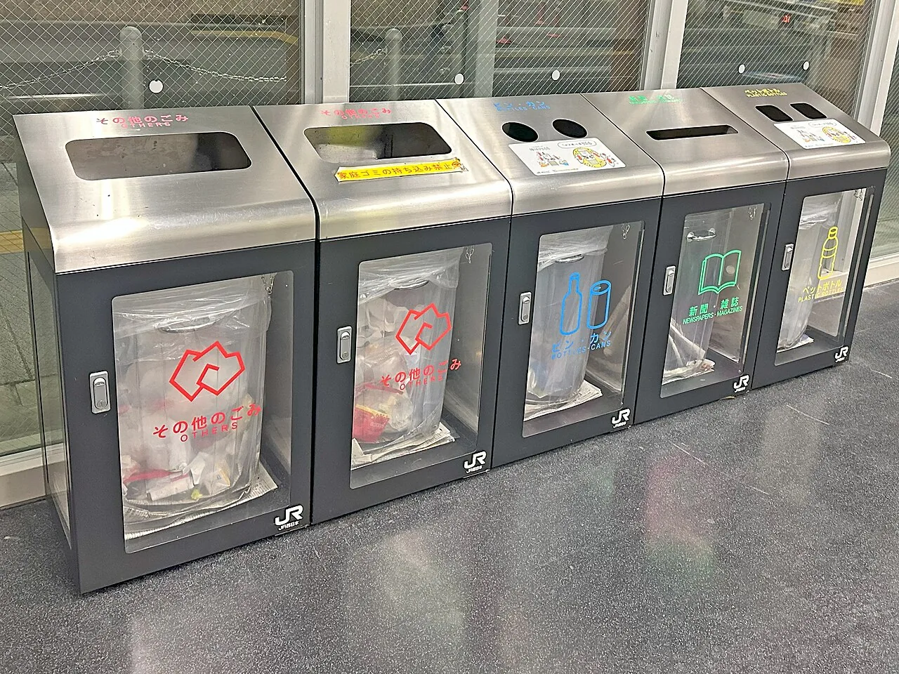 Five sorted waste containers standing in a row on a Japanese station concourse, each with a clear panel
