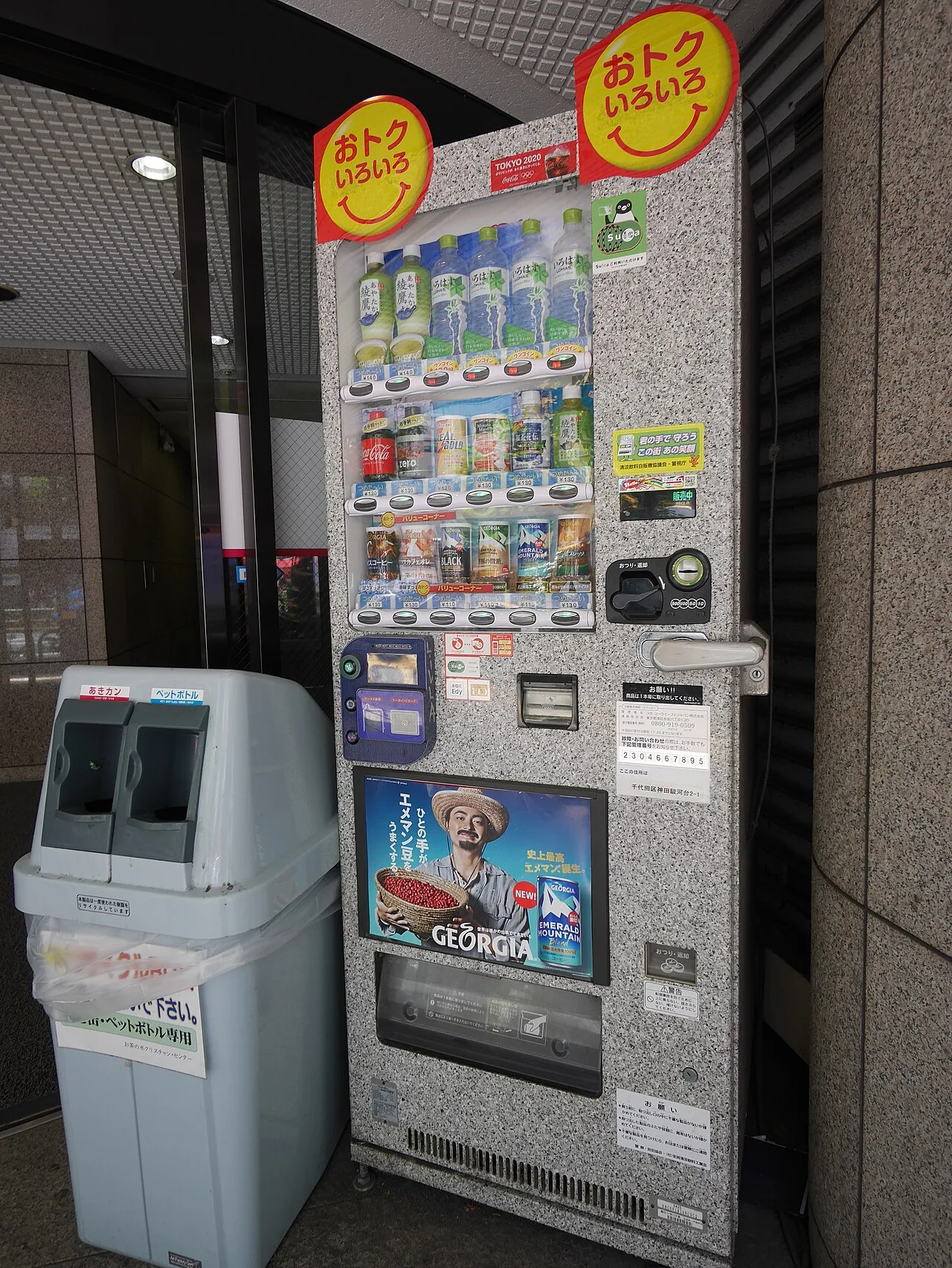 Two drink vending machines against a wall with a separate collection box standing beside them