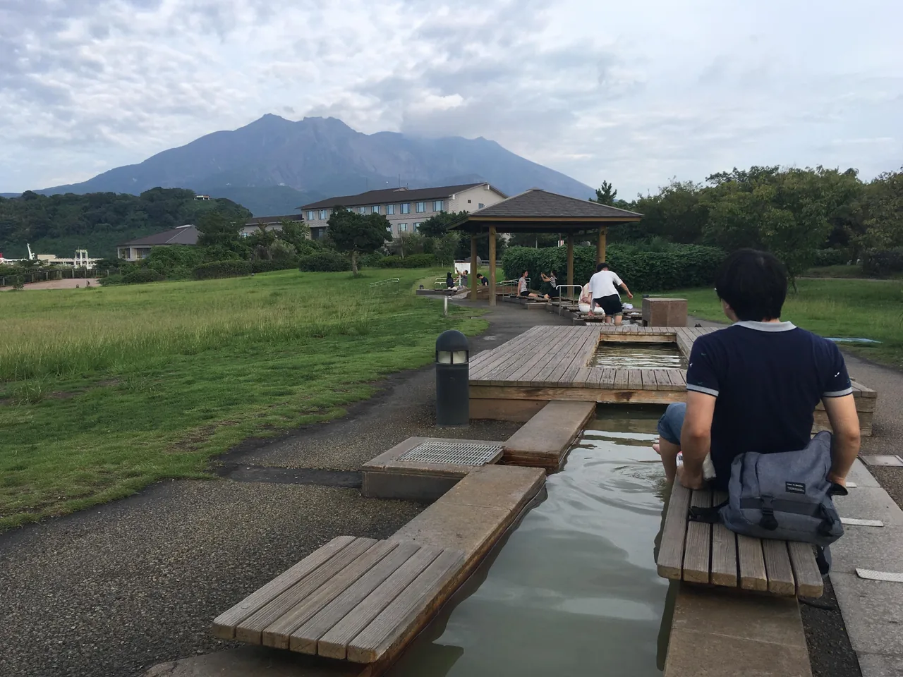 A long wooden foot bath in the lava park with people sitting along it and Sakurajima behind
