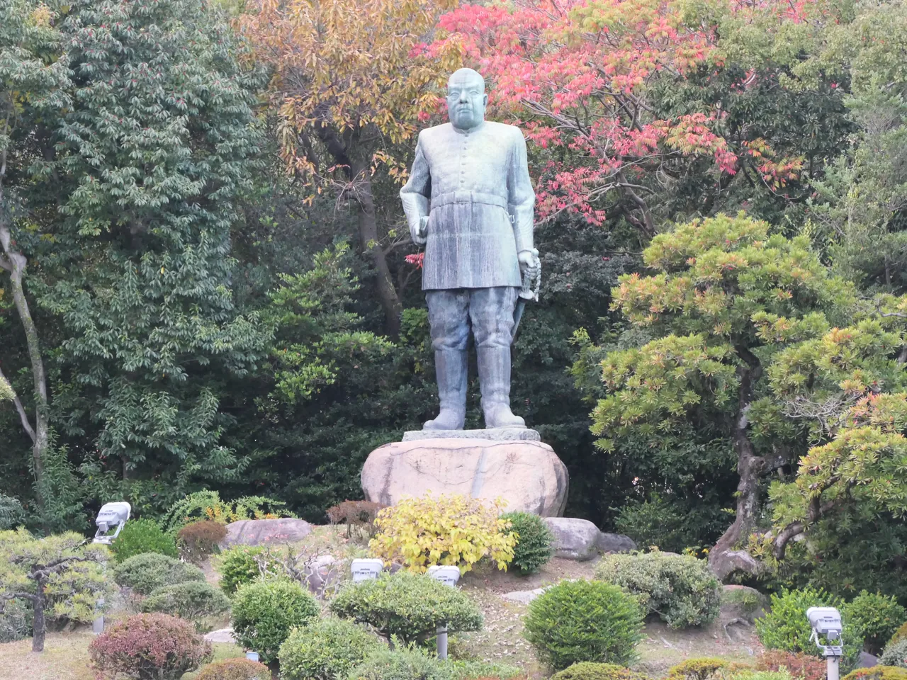 The bronze statue of Saigo Takamori in uniform on a stone base, with pink azaleas and clipped greenery around it