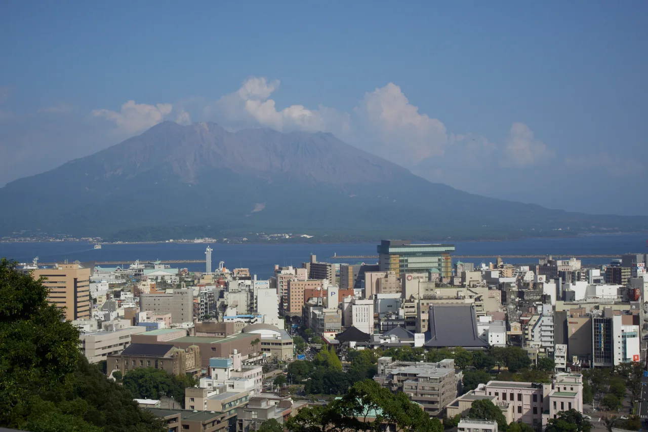 Sakurajima standing across Kinko Bay under a blue sky, with the buildings of central Kagoshima filling the foreground
