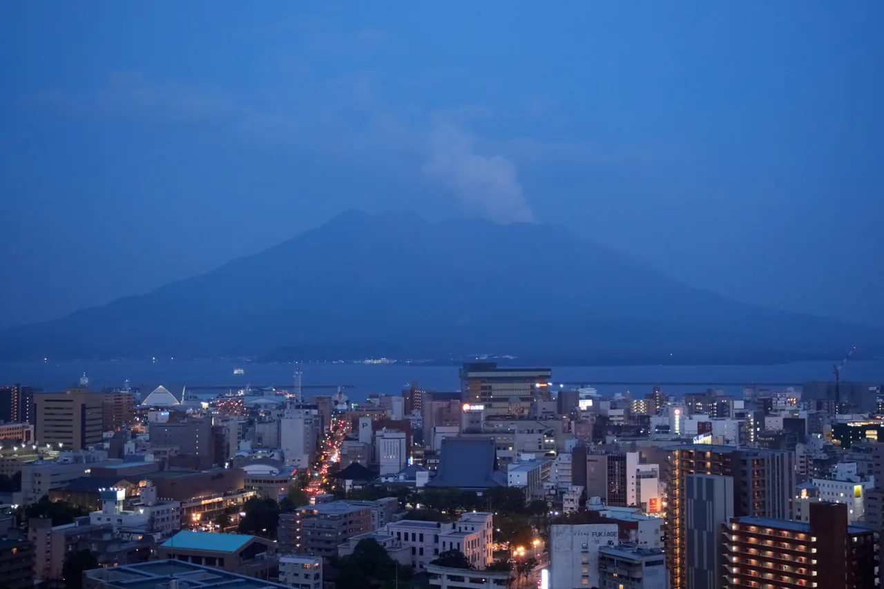 Central Kagoshima lit up at dusk, with Sakurajima rising across the bay behind it