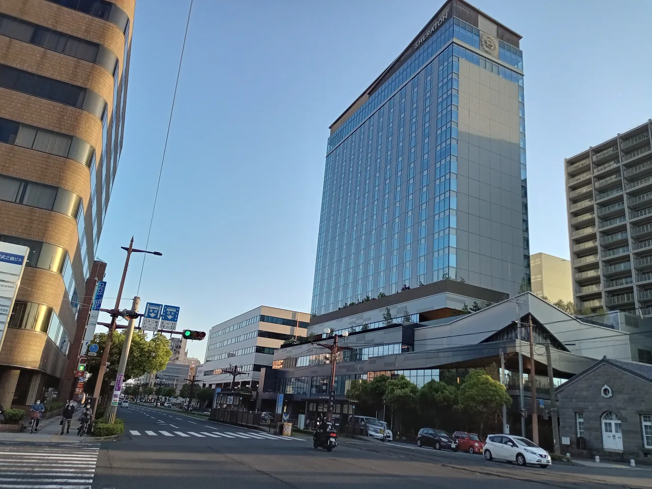 A glass-fronted high-rise hotel standing beside a city crossing in Kagoshima, with traffic lights and low buildings in front of it