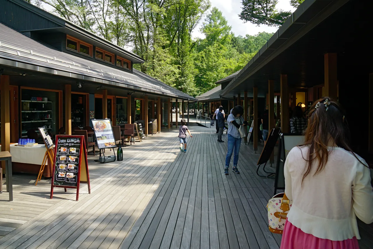 A wooden boardwalk between low shop buildings under tall trees