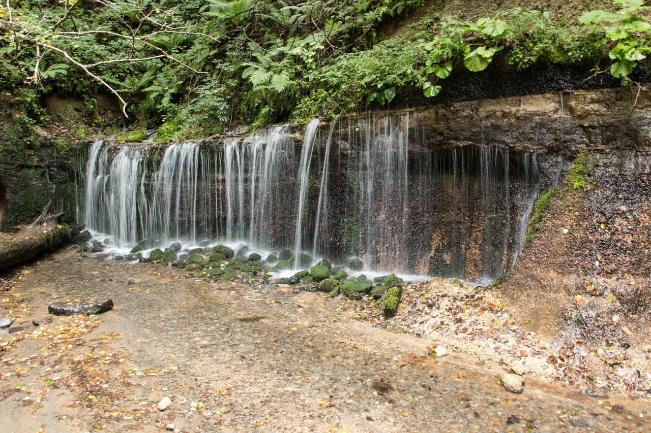 A wide low curtain of water running down a dark rock face into a shallow pool