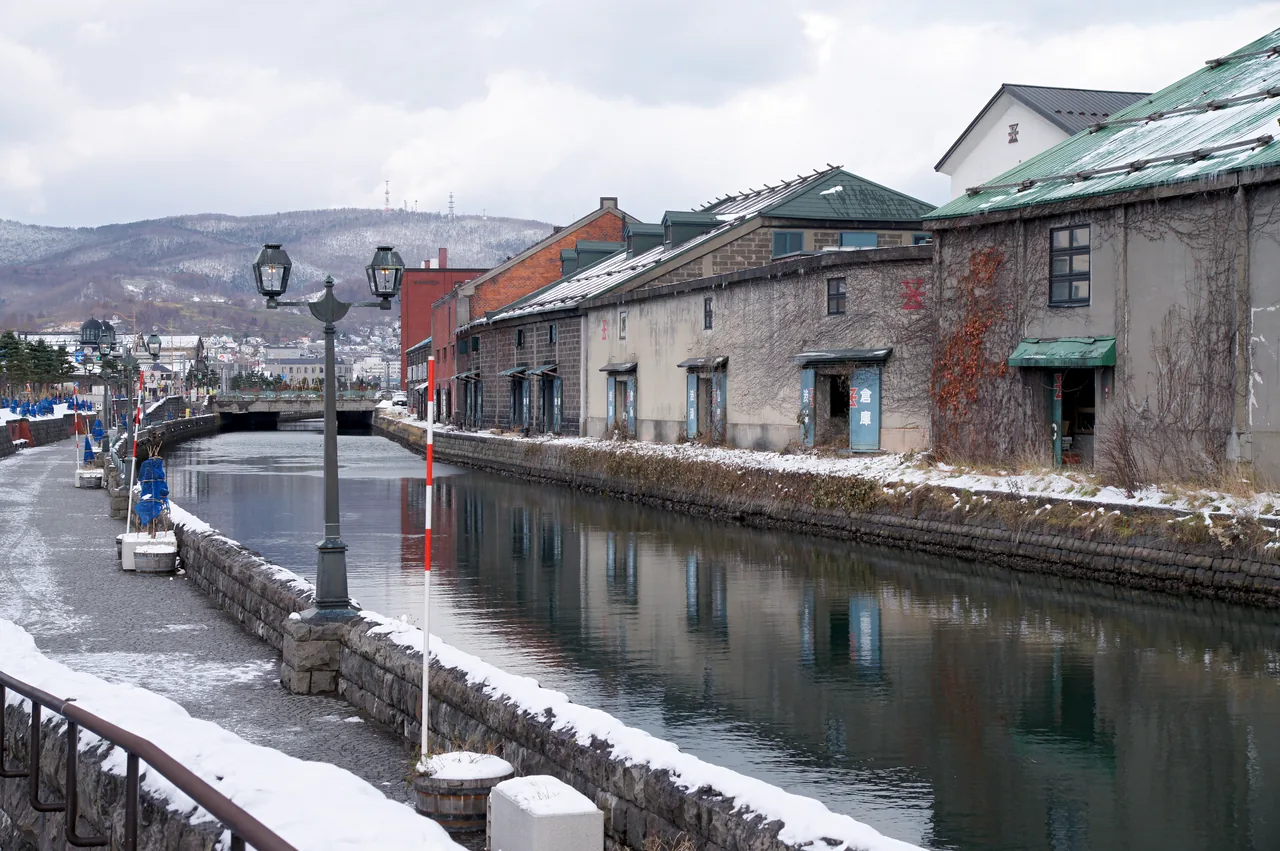 Snow-covered canal walkway with gas lamps and warehouses across the water on a grey winter day