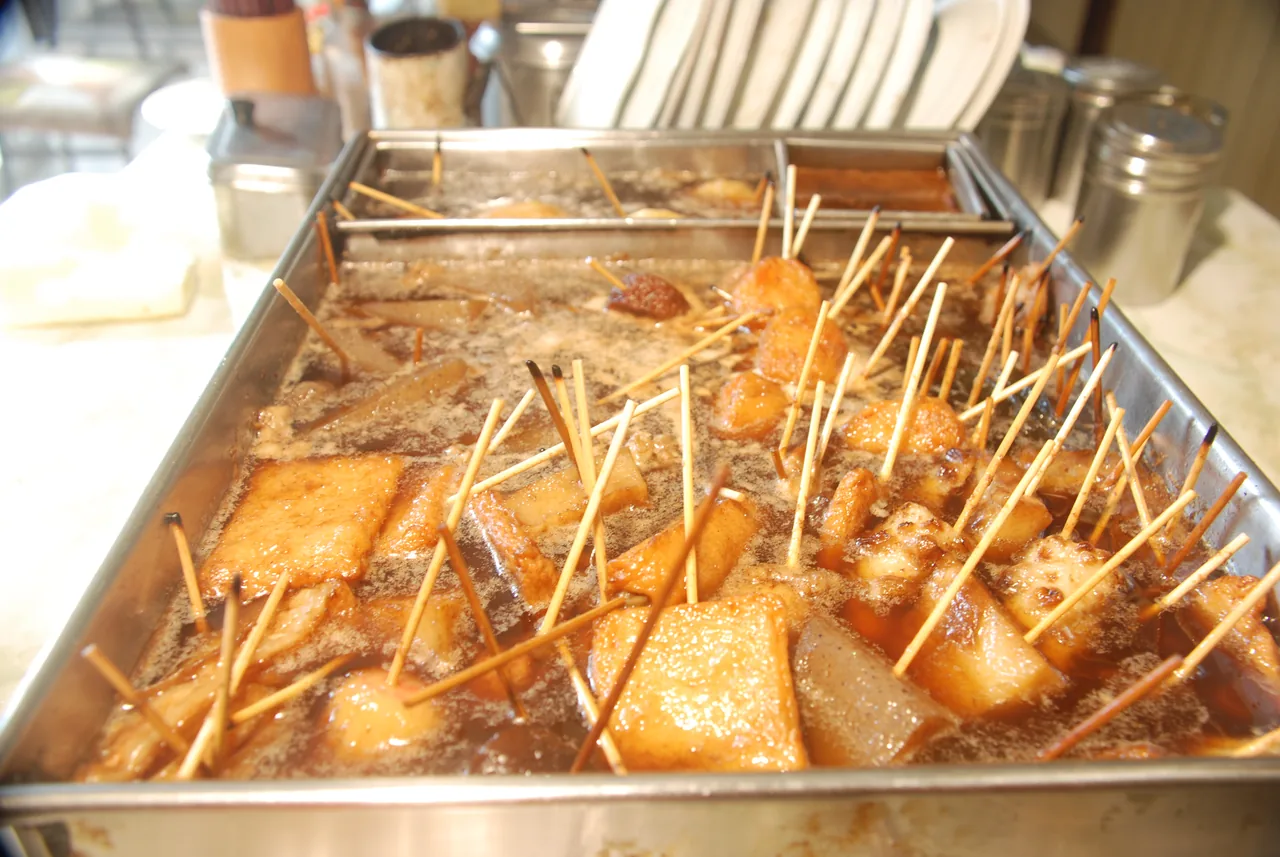 A metal tray of dark oden broth with wooden skewers standing in it