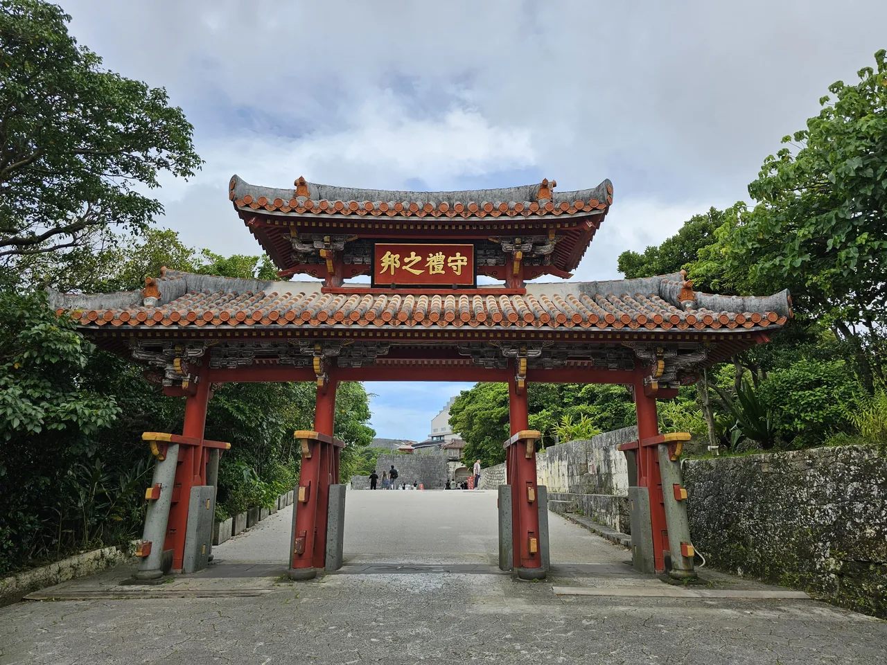 The red-tiled Shureimon gate with its painted plaque, standing among trees below Shuri Castle