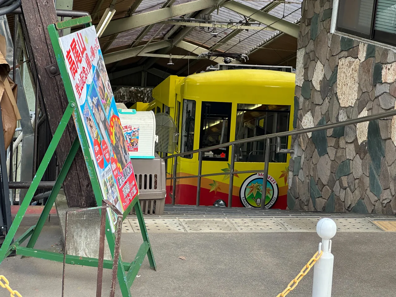 A yellow cable car waiting at the Mt Takao station platform beside a noticeboard