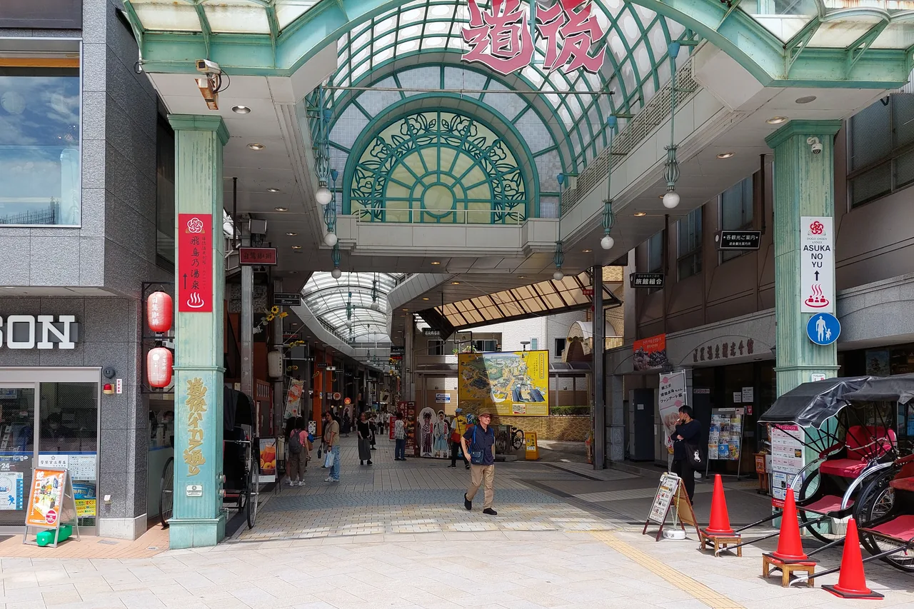Entrance to the Dogo shopping arcade, a green iron-and-glass arch marked 道後, with a few people and a rickshaw