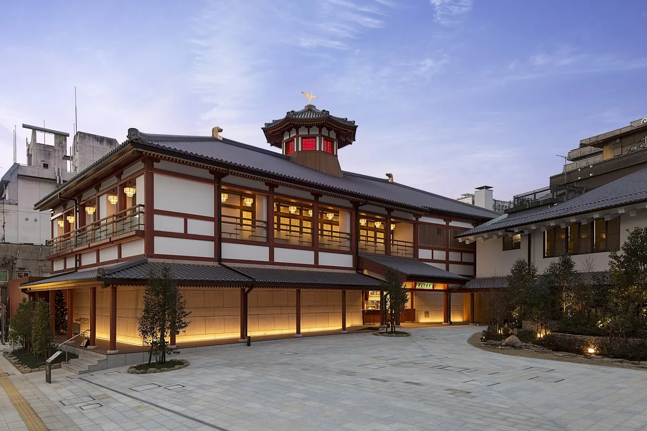 Asuka-no-Yu lit up at dusk, with white walls, red pillars and a tower on the roof, and an empty stone courtyard in front