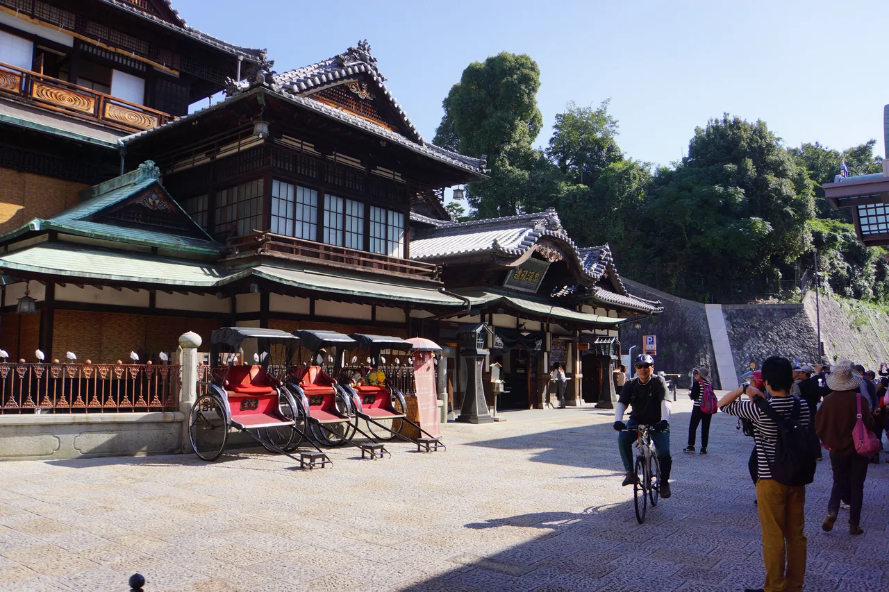 Dogo Onsen Honkan seen from the side on a clear day, a row of red rickshaws parked in front and a few visitors