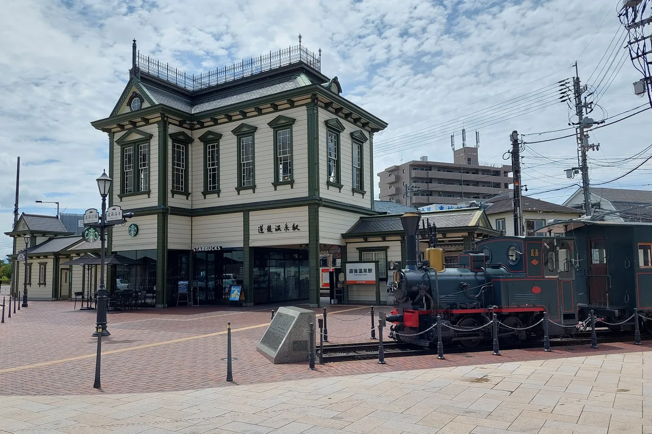 The Western-style wooden Dogo Onsen Station building with a Botchan Train, a steam-style engine and carriage, standing in front on a cloudy day