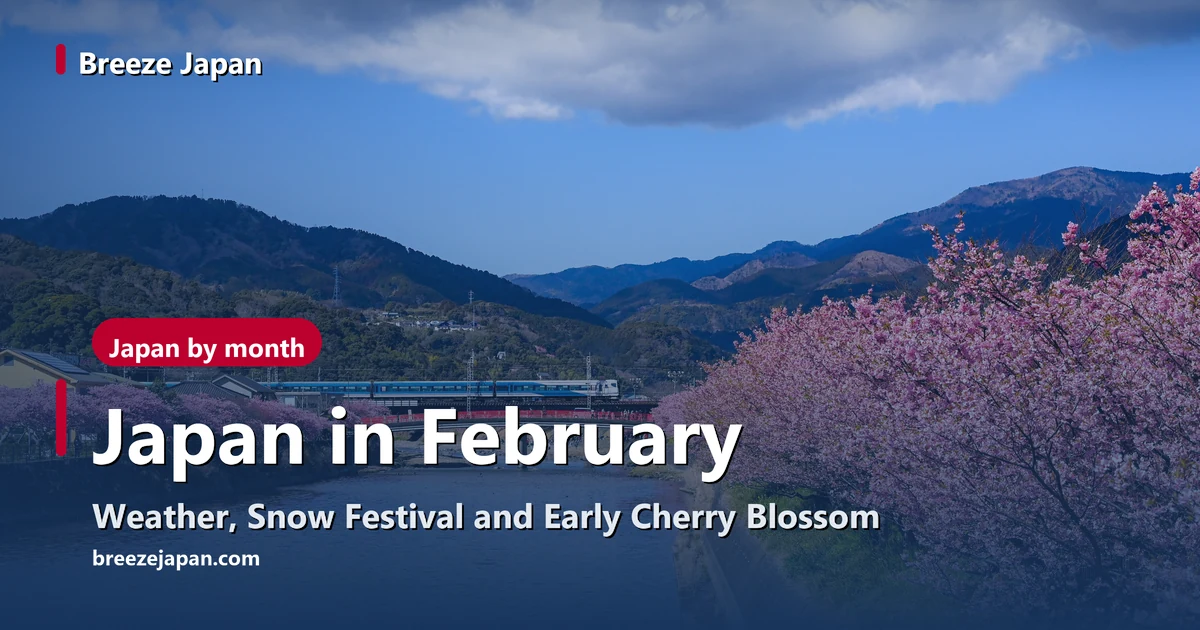 Pink Kawazu cherry trees along both banks of a river and a white and blue limited express train crossing a bridge, with blue sky and hills