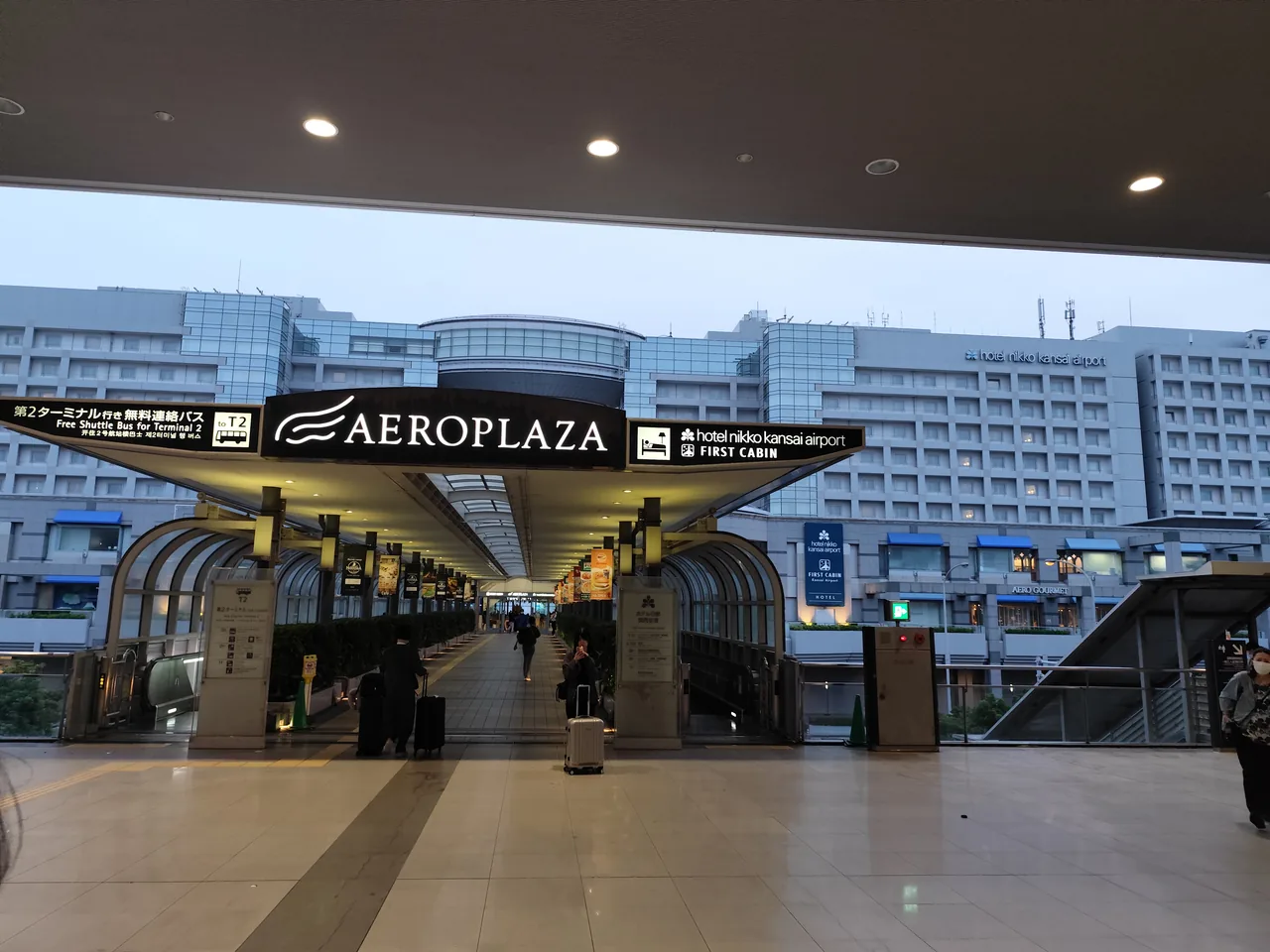 Covered walkway to the Aeroplaza at dusk with Aeroplaza, Hotel Nikko and First Cabin signs and the hotel building behind