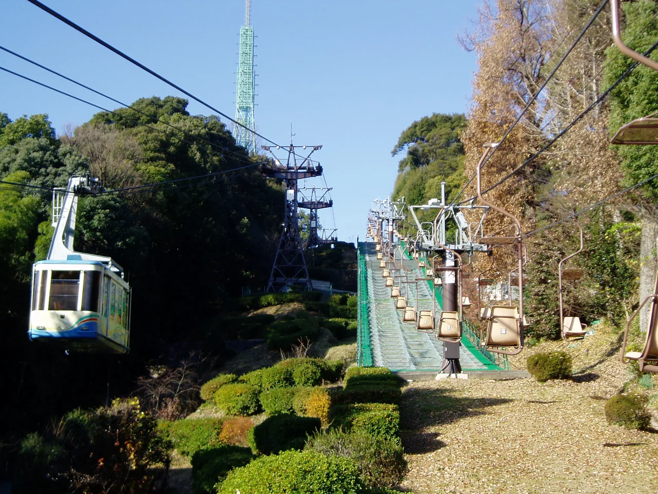 A ropeway gondola on the left climbing a wooded slope beside a line of chairlift seats