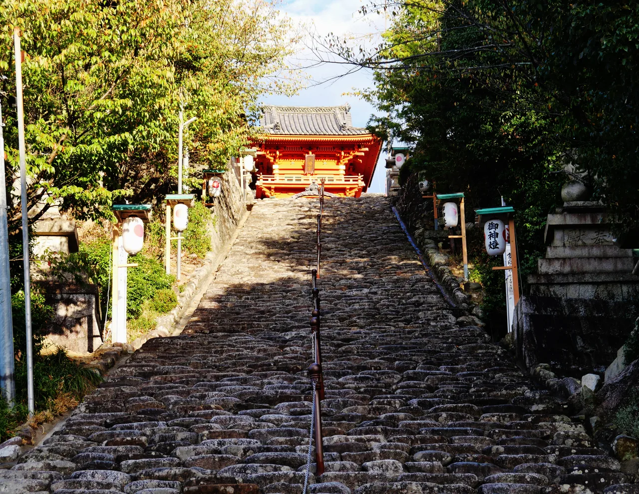 A long, straight flight of stone steps between trees leading up to the red gate of Isaniwa Shrine