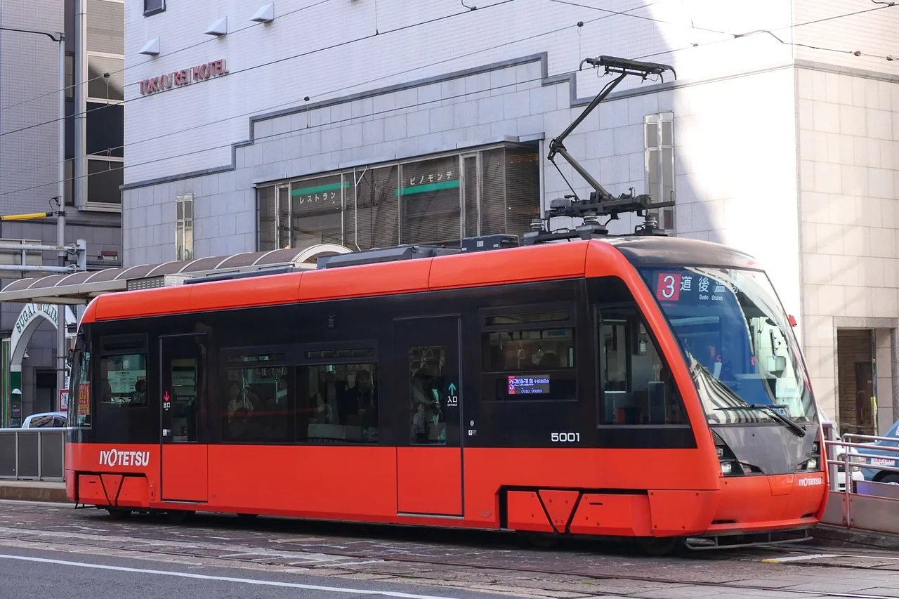 A red low-floor Iyotetsu tram on a street in front of a hotel, its destination sign reading 3 道後温泉